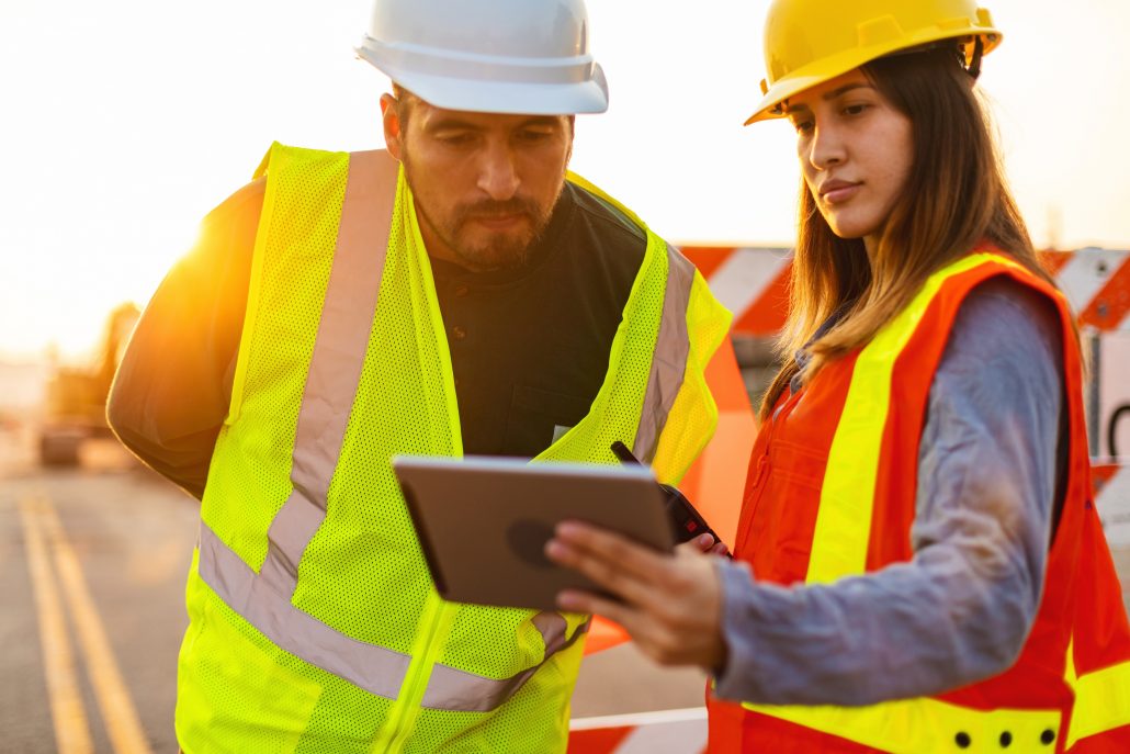 Dos profesionales del mantenimiento de pavimentos, que llevan cascos y chalecos de seguridad de alta visibilidad, consultan una tableta mientras planifican los trabajos de mantenimiento de carreteras y reparación del asfalto.